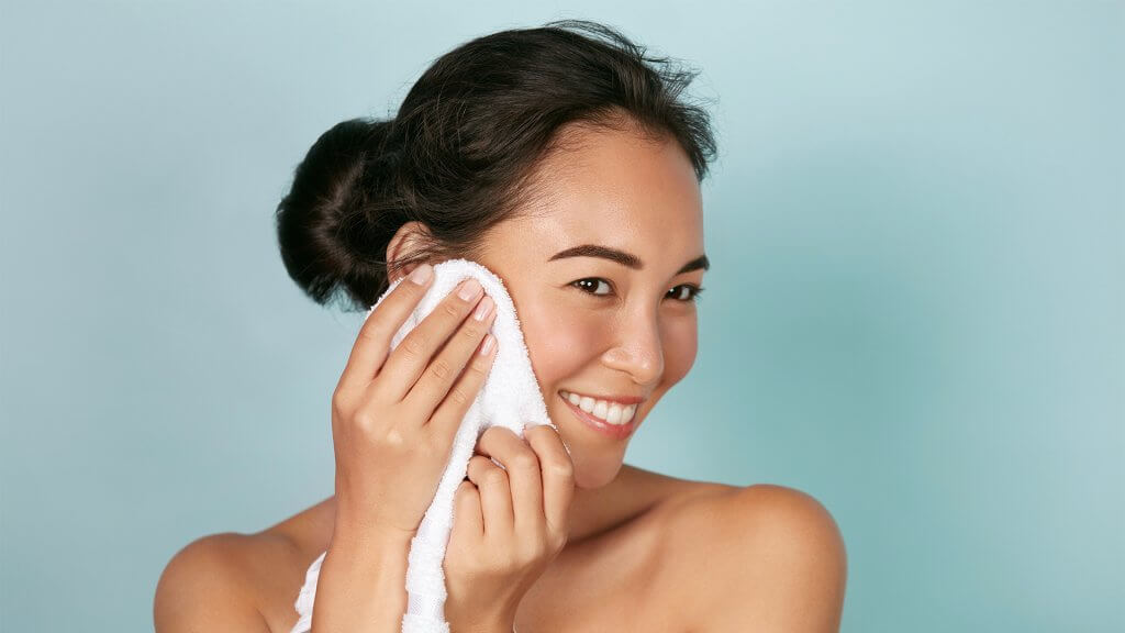 Woman gently dabbing her face with a white towel, against a blue background